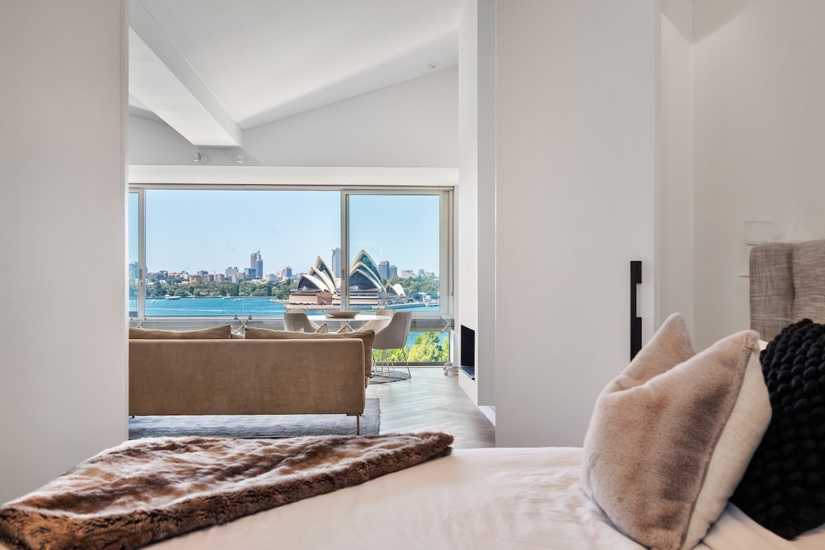 The bedroom features a spacious bed with neutral linens, complemented by decorative pillows. A cozy throw is draped across the bed. Large windows offer a stunning view of Sydney Harbour and the iconic Opera House, filling the room with natural light.