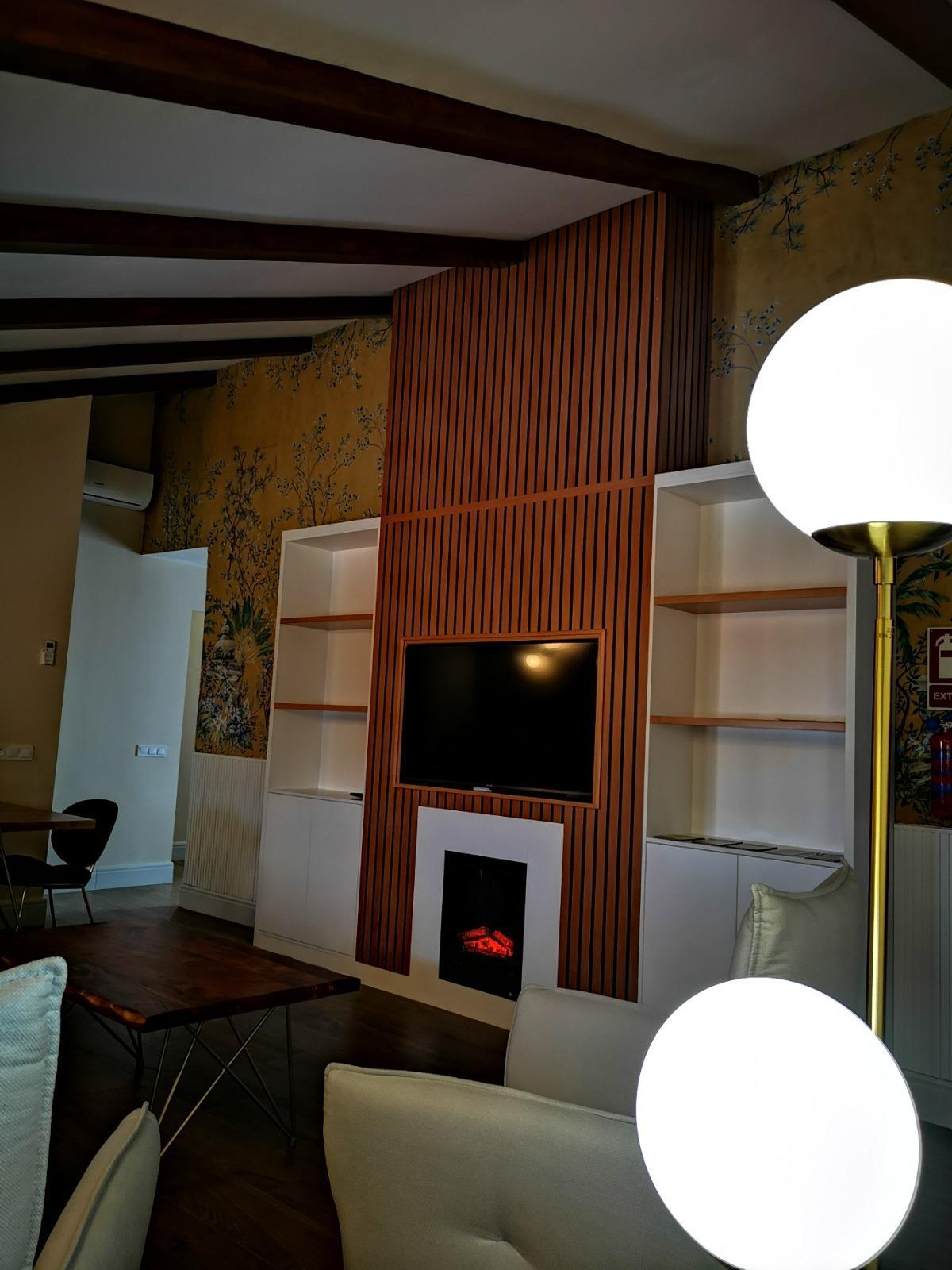 A living area is presented with stylish wooden accents and a mounted television above a modern electric fireplace. Soft white light from a floor lamp illuminates the space, which features a wooden coffee table and plush seating. Decorative wallpaper enhances the overall ambiance.