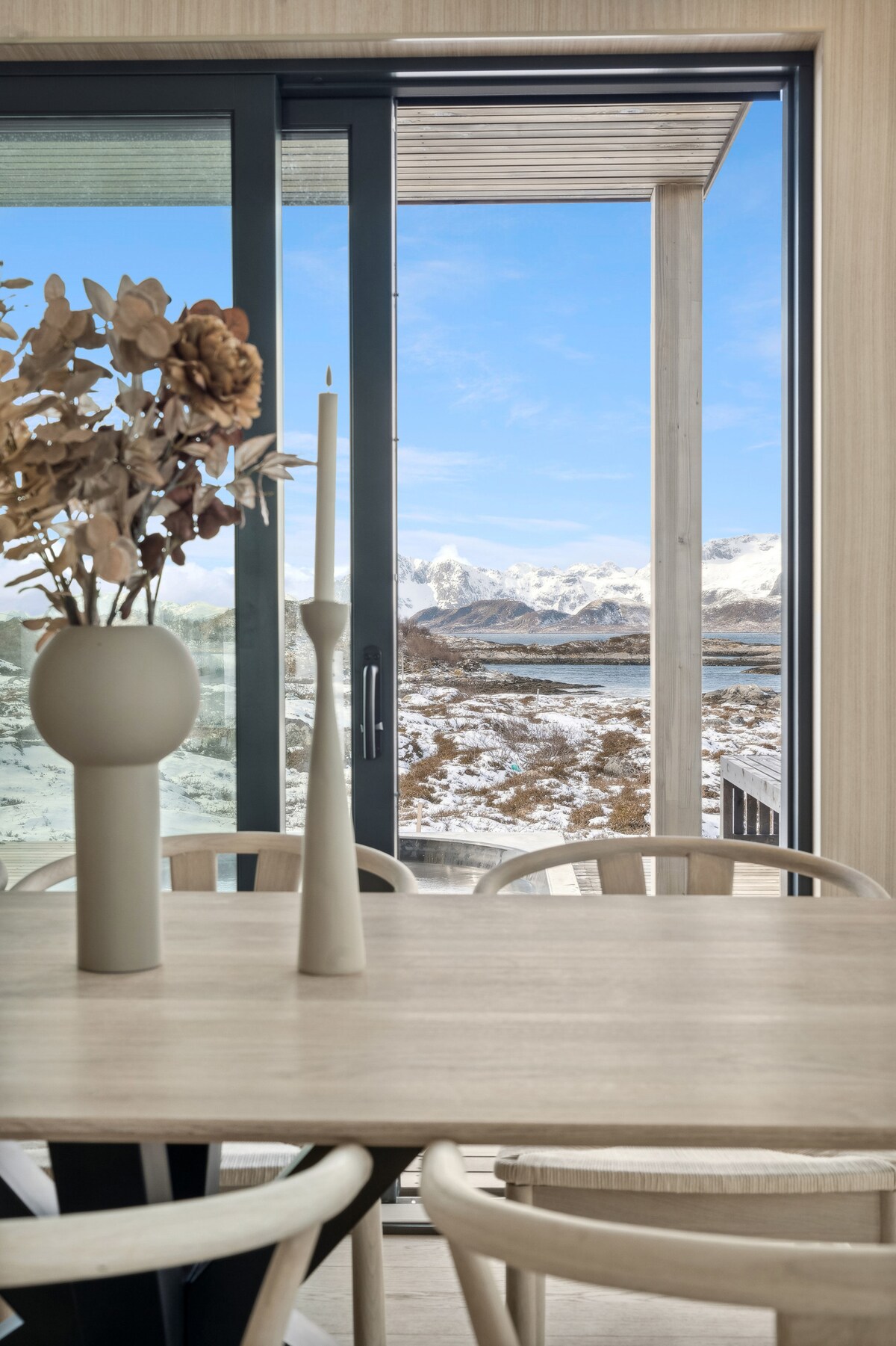 A dining area features a light wooden table surrounded by four chairs. A decorative vase with dried flowers and a candle holder are placed on the table. Glass doors open to a snowy landscape with mountains in the background and a clear blue sky.
