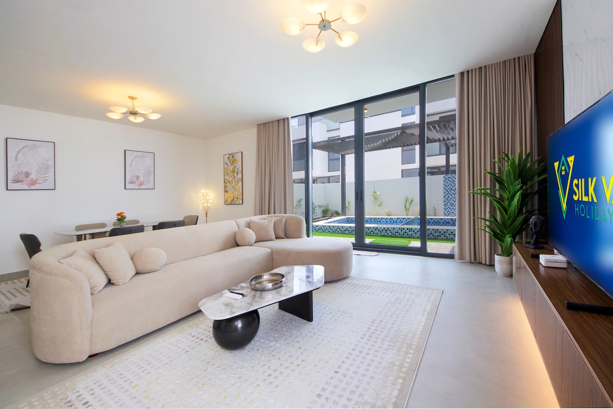 A contemporary living space features a curved, light-colored sofa with plush cushions, positioned near large windows that open to a private garden. A stylish coffee table sits in front of the sofa, while a flat-screen TV is mounted on a wooden cabinet. Dining area is visible in the background.