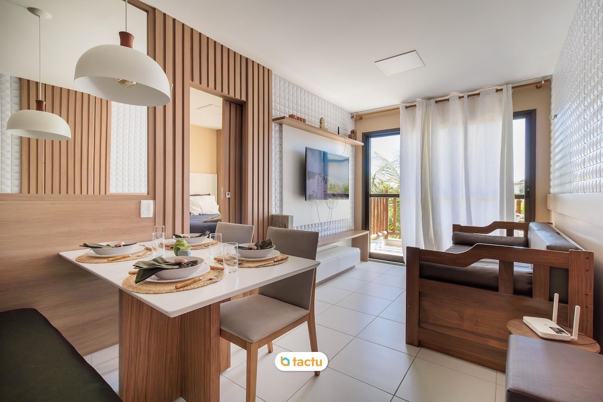 The living area features a modern dining table set for four and a comfortable seating area with wooden accents. A large wall-mounted TV is visible, along with a glass door leading to a balcony. Soft natural light illuminates the space, enhancing a relaxed atmosphere.