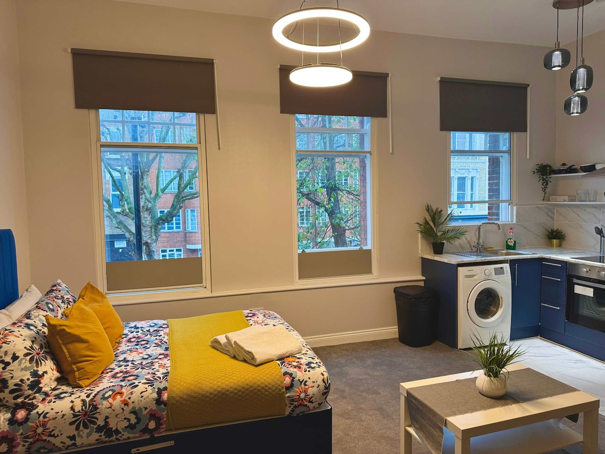 A studio space features a standard double bed with a floral design and a yellow throw. Natural light enters through three windows, illuminating the cozy area. A small table with a potted plant and a modern kitchen with a washer are visible in the background.