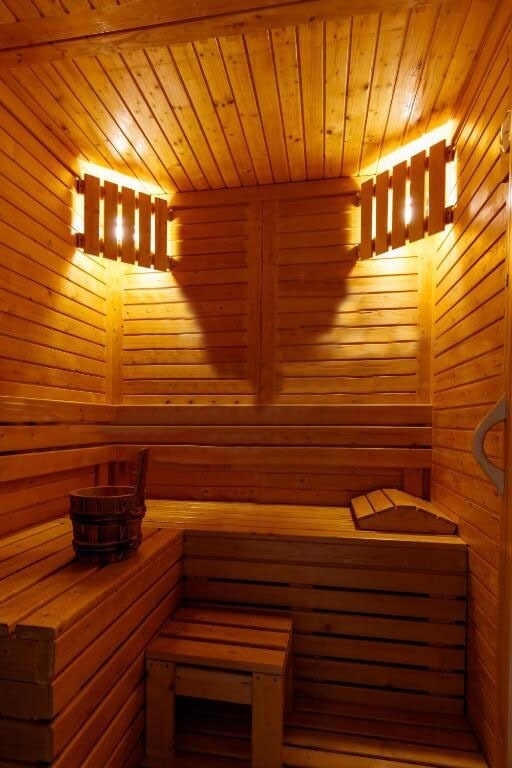 A sauna area is presented, featuring warm wooden paneling and built-in benches. Soft lighting enhances the ambiance, creating a soothing environment. A wooden bucket and a small stool are placed within the space, inviting relaxation.