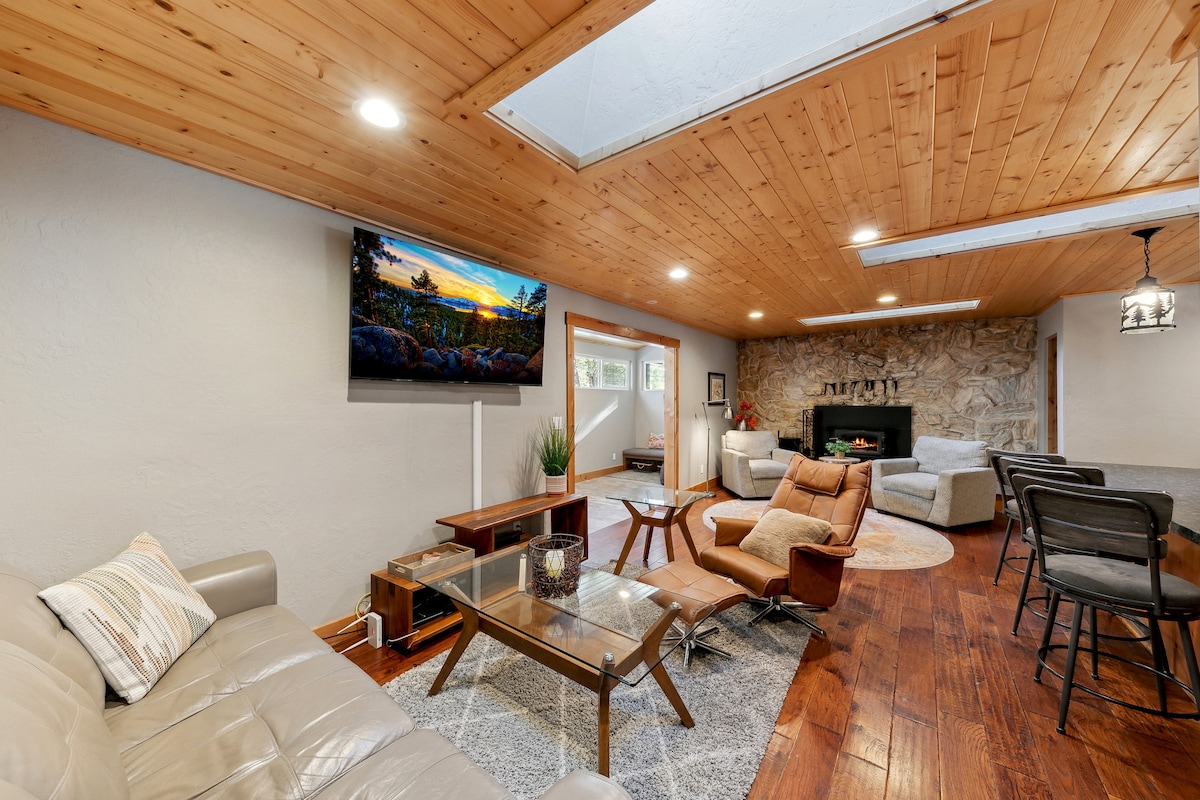 A cozy living area features polished wood flooring and accent wood paneling on the ceiling. A large sectional sofa is paired with a reclining chair, and a glass coffee table is centered on a soft area rug. A wall-mounted TV is visible above a stone fireplace.