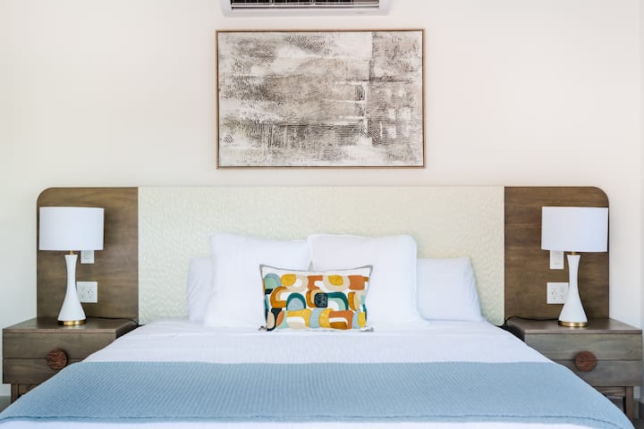 - Светла и весела соба со мека постелнина и разиграна перница со акцент
- Топлите дрвени тонови се среќаваат со свежа бела боја за изглед кој е удобен и полиран
- Центрираните апстрактни уметнички дела додаваат карактер на овој мирен простор за спиење