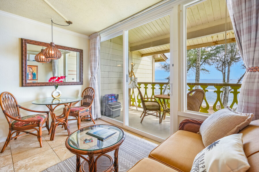 A cozy seating area features a light-colored sofa and a glass coffee table. Two wicker chairs surround a glass dining table, set against expansive sliding glass doors that provide views of the ocean. Soft curtains frame the doorway, enhancing the coastal ambiance.
