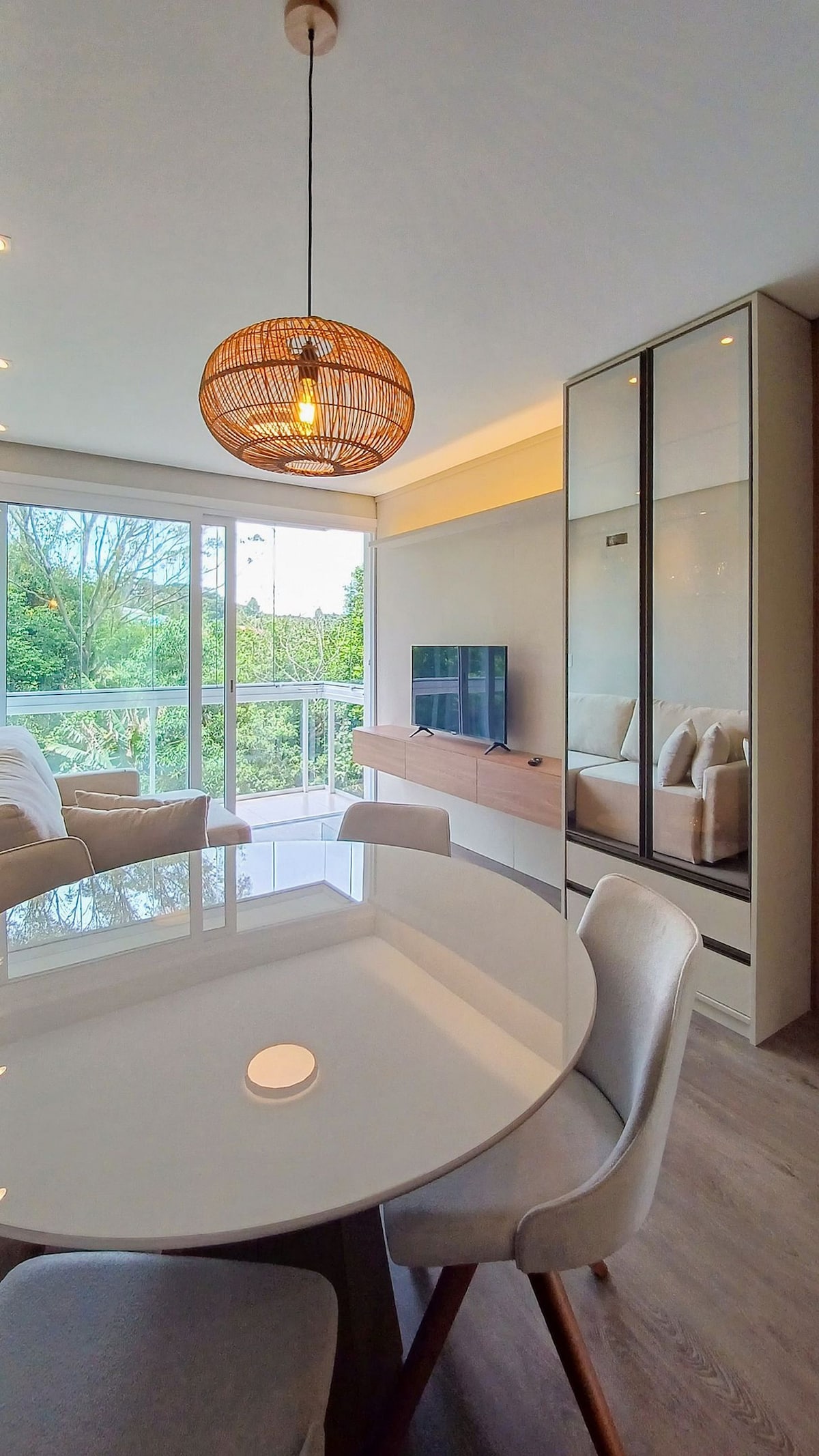 A dining area features a round table with light-colored chairs, complemented by a decorative pendant light overhead. A large window offers views of the greenery outside, while a stylish TV unit is visible in the background, enhancing the modern ambiance of the space.