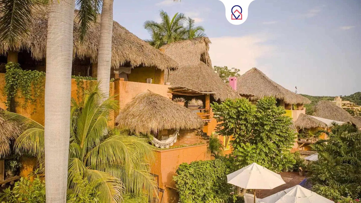 The image captures a vibrant complex of thatched-roof buildings nestled among lush greenery. Palm trees and tropical plants surround the structures, while sun umbrellas are visible near a shared pool area, inviting relaxation in a scenic environment.