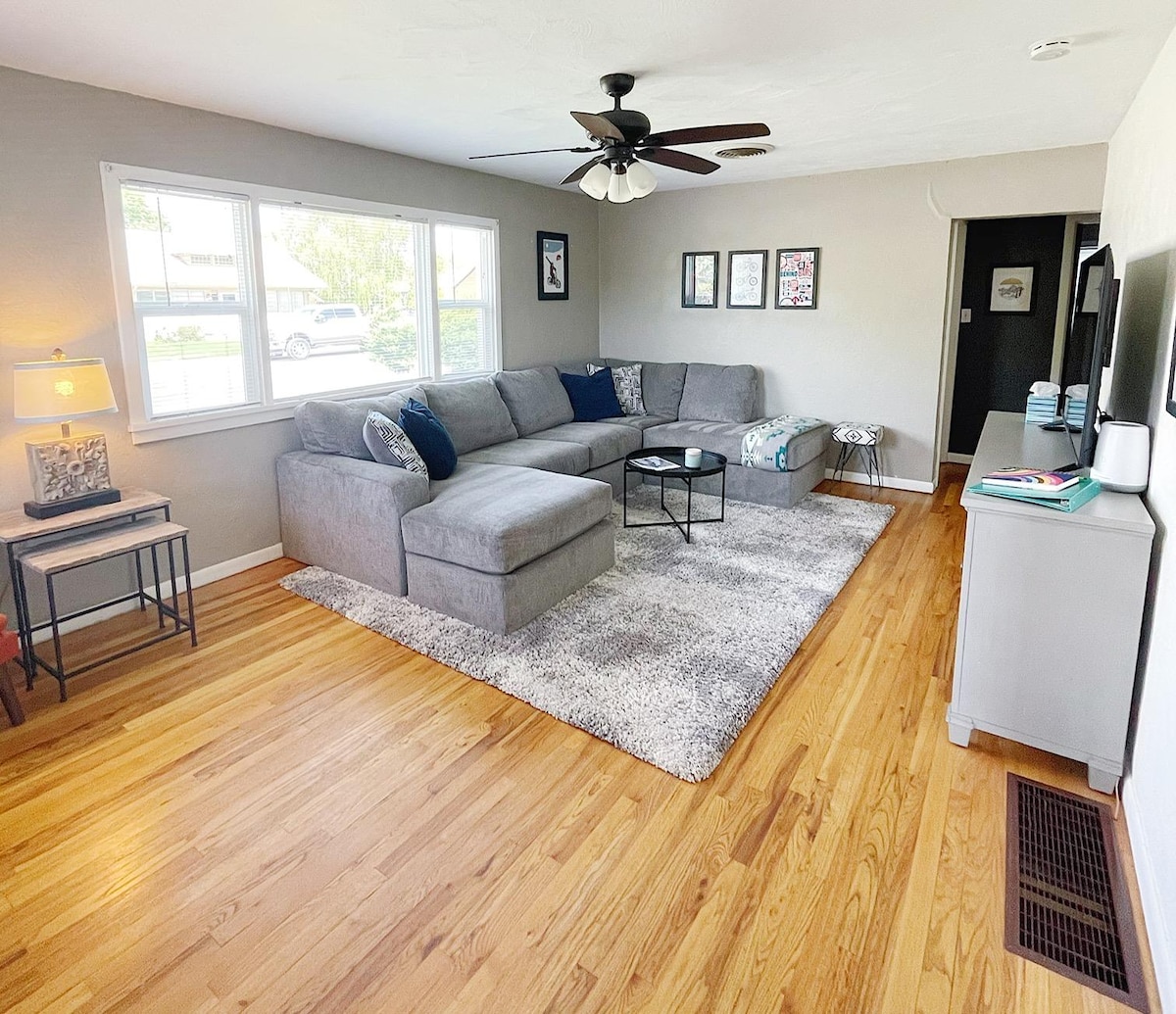 A bright living space features a large sectional sofa complemented by a decorative side table, all arranged on a plush area rug. Natural light floods through multiple windows, illuminating the hardwood floor and surrounding artwork on the walls. A flat-screen TV can be seen on the cabinet.