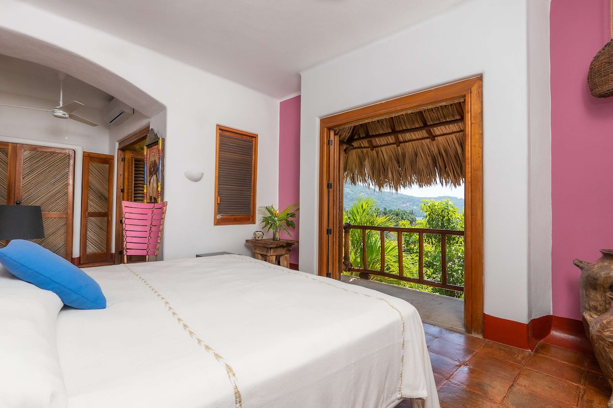 A cozy bedroom is showcased, featuring a spacious bed with crisp white linens and a blue accent pillow. Natural light floods the room from an open door leading to a private terrace, adorned with tropical foliage and a thatched roof, enhancing the serene atmosphere.