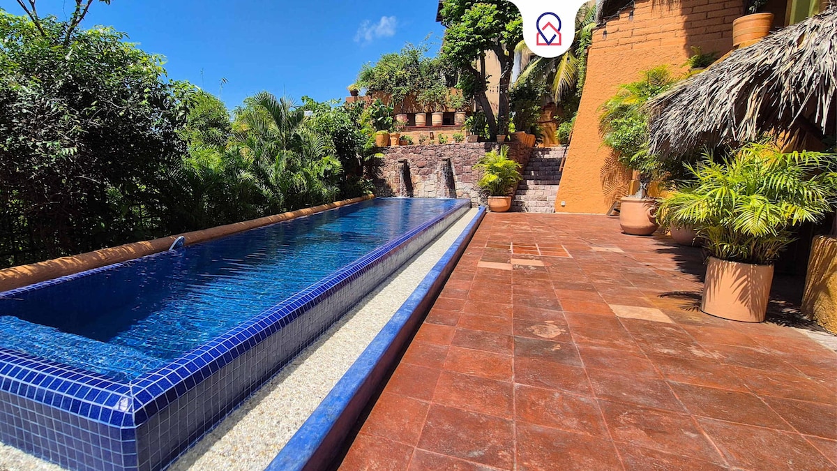 A long, inviting swimming pool is bordered by a smooth stone pathway, surrounded by lush greenery and terracotta planters. Palm trees and vibrant shrubs create a serene environment, while the clear blue sky adds brightness to the scene.