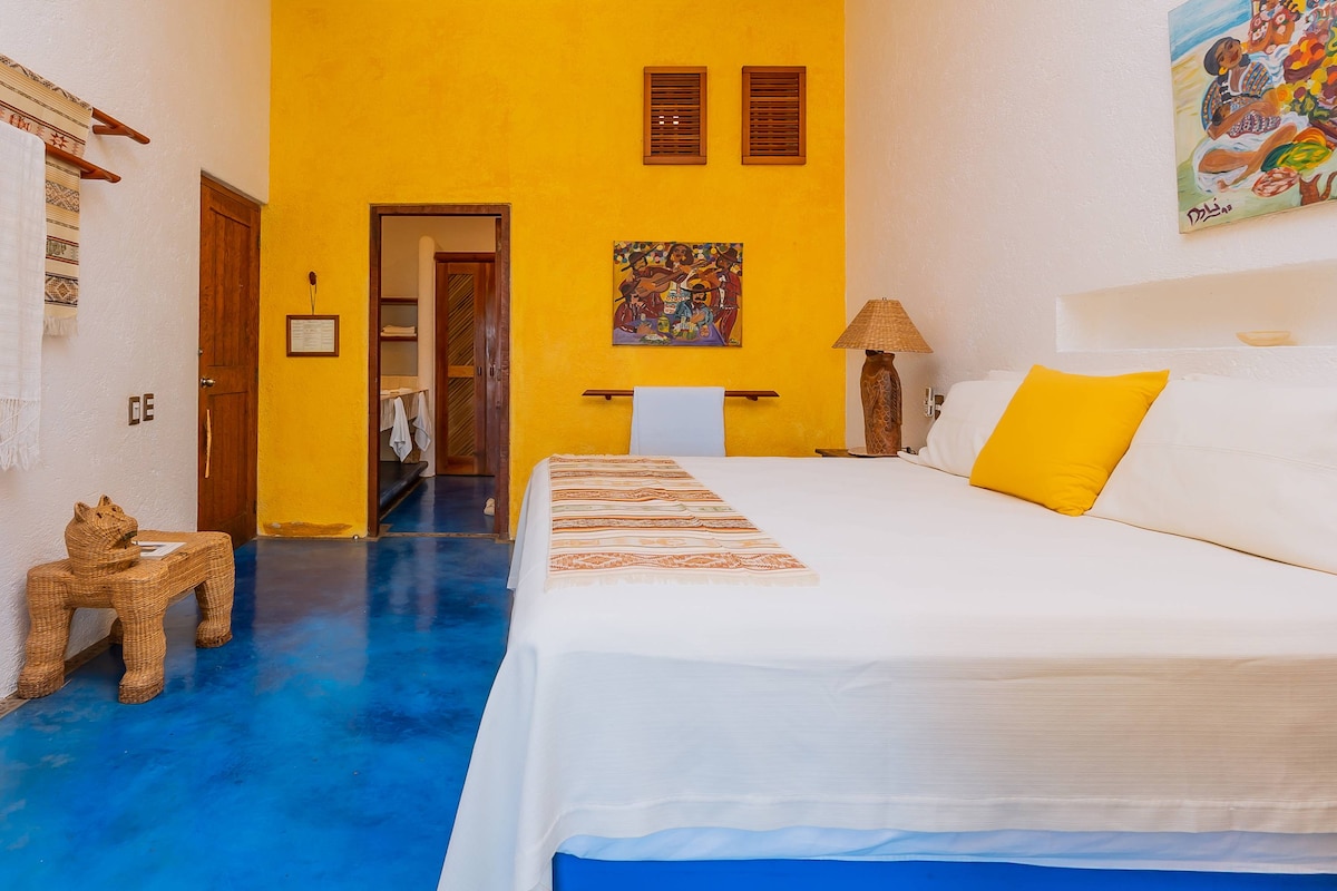 The studio features a bright yellow accent wall and a spacious bed with white linens. A woven table and lamp add texture to the room. Colorful wall art and wooden shutters enhance the inviting atmosphere, while a glimpse of the bathroom is visible through an open door.