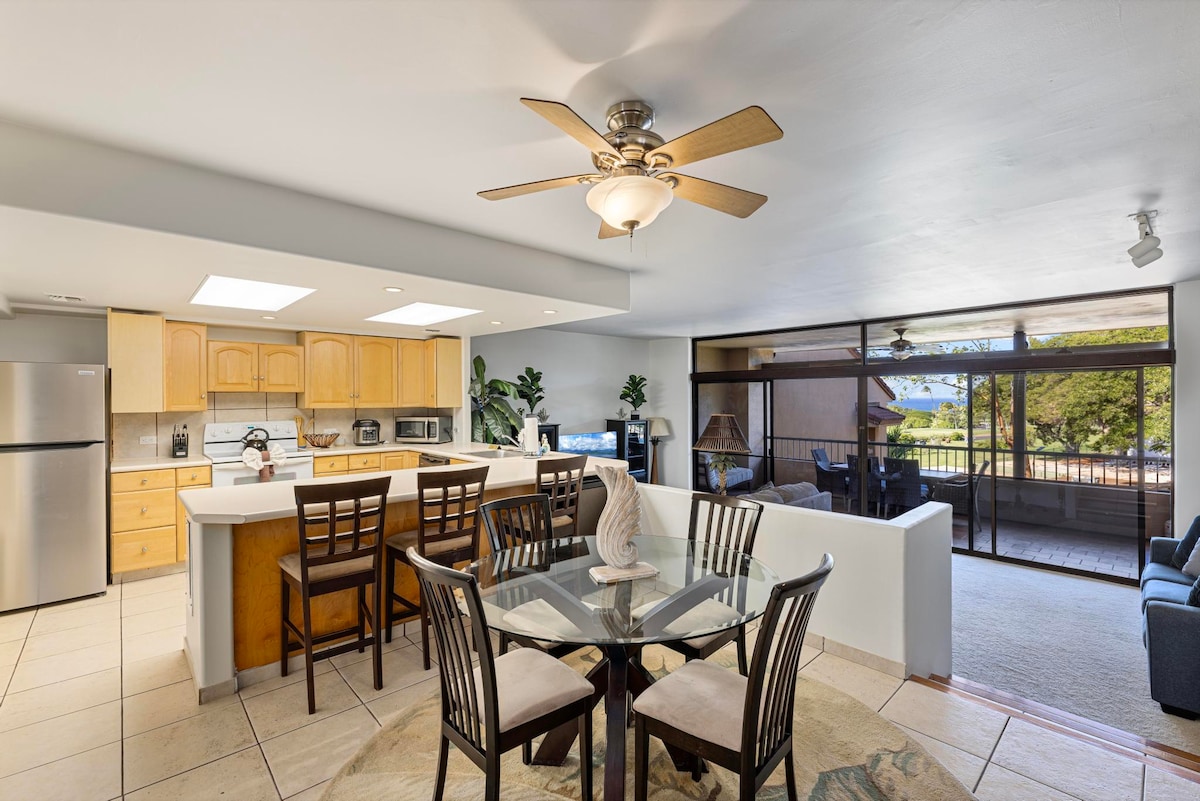 An open-concept kitchen and dining area features light wood cabinets and a spacious island with seating. A glass dining table is surrounded by six chairs, illuminated by a ceiling fan. Large sliding glass doors connect to the outdoor space, offering views of the lush surroundings.