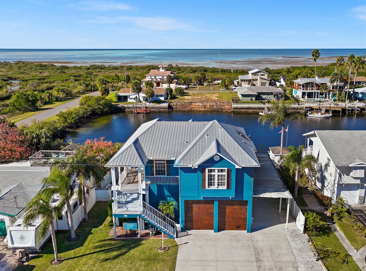 An aerial view showcases a colorful stilt home adjacent to saltwater canals. The property features a spacious driveway and easy access to the water, with lush greenery and nearby homes visible in the background. The Gulf of Mexico glistens in the distance.