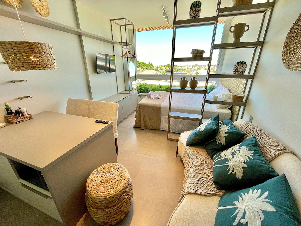 The open-plan living area features a comfortable sofa adorned with decorative cushions and a light dining table. A spacious bed with neutral bedding is positioned near a window, allowing natural light to fill the room. Decorative plant baskets enhance the cozy atmosphere.