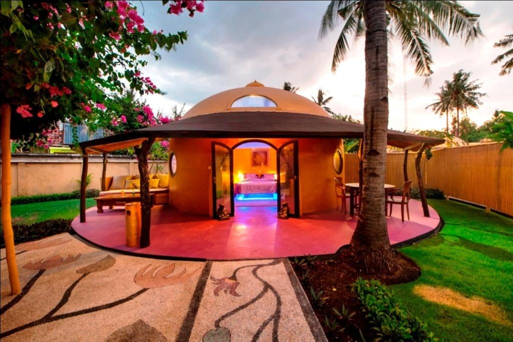 A round structure with a thatched roof is surrounded by lush greenery and colorful flowers. The entrance features circular doorways opening to a softly lit interior with ambient blue lighting. Outdoor seating areas are positioned on a vibrant patio, inviting relaxation amidst the natural surroundings.