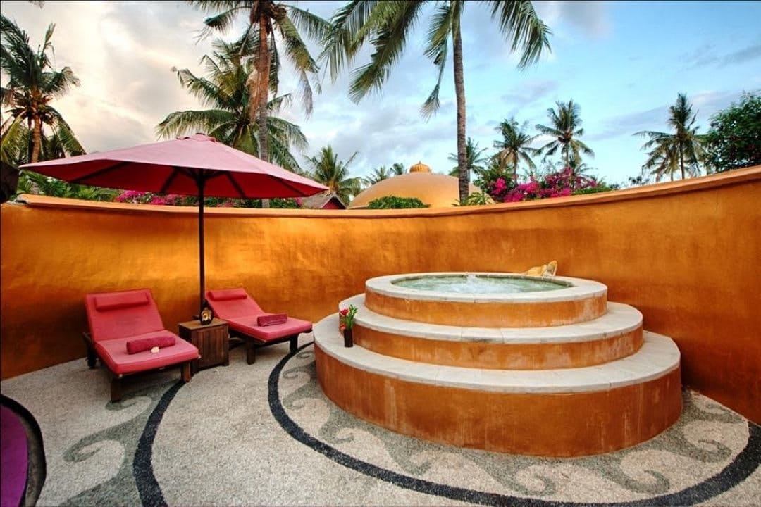 A private outdoor area features a circular hot tub, surrounded by tiered stone steps. Two lounge chairs with matching cushions sit beneath a large umbrella, while vibrant tropical plants line a bright orange wall. The scene is set against a backdrop of palm trees and a blue sky.
