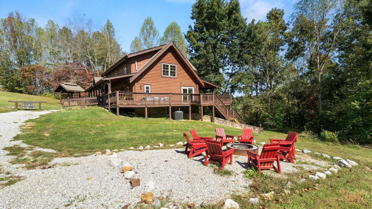 A rustic cabin sits on a grassy area surrounded by trees, featuring a spacious deck and multiple outdoor seating areas. A fire pit is centrally located, encircled by six red Adirondack chairs. Gravel paths and natural landscaping enhance the outdoor setting.