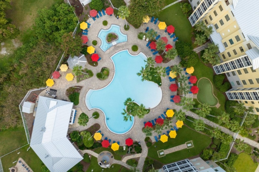 An overhead view captures the resort's pool area, featuring a large, irregularly shaped pool surrounded by vibrant umbrellas in red and yellow. Lush landscaping and palm trees enhance the atmosphere, with lounge chairs and a smaller putting green visible in the vicinity.