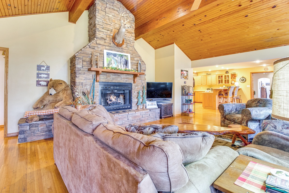A spacious living area features a stone fireplace as a focal point, surrounded by comfortable seating and wood detailing. Large windows allow light to fill the space, enhancing the natural warm tones of the wooden ceiling and flooring.