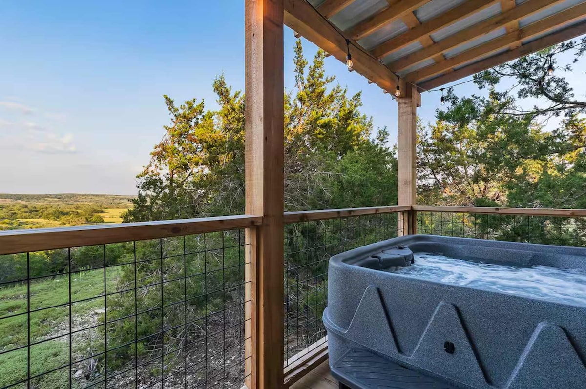 A covered outdoor space features a private hot tub, inviting relaxation with views of the surrounding lush greenery. The wooden structure showcases a rustic design, with railing framing the area. Natural light filters in through the open sky, enhancing the serene atmosphere.