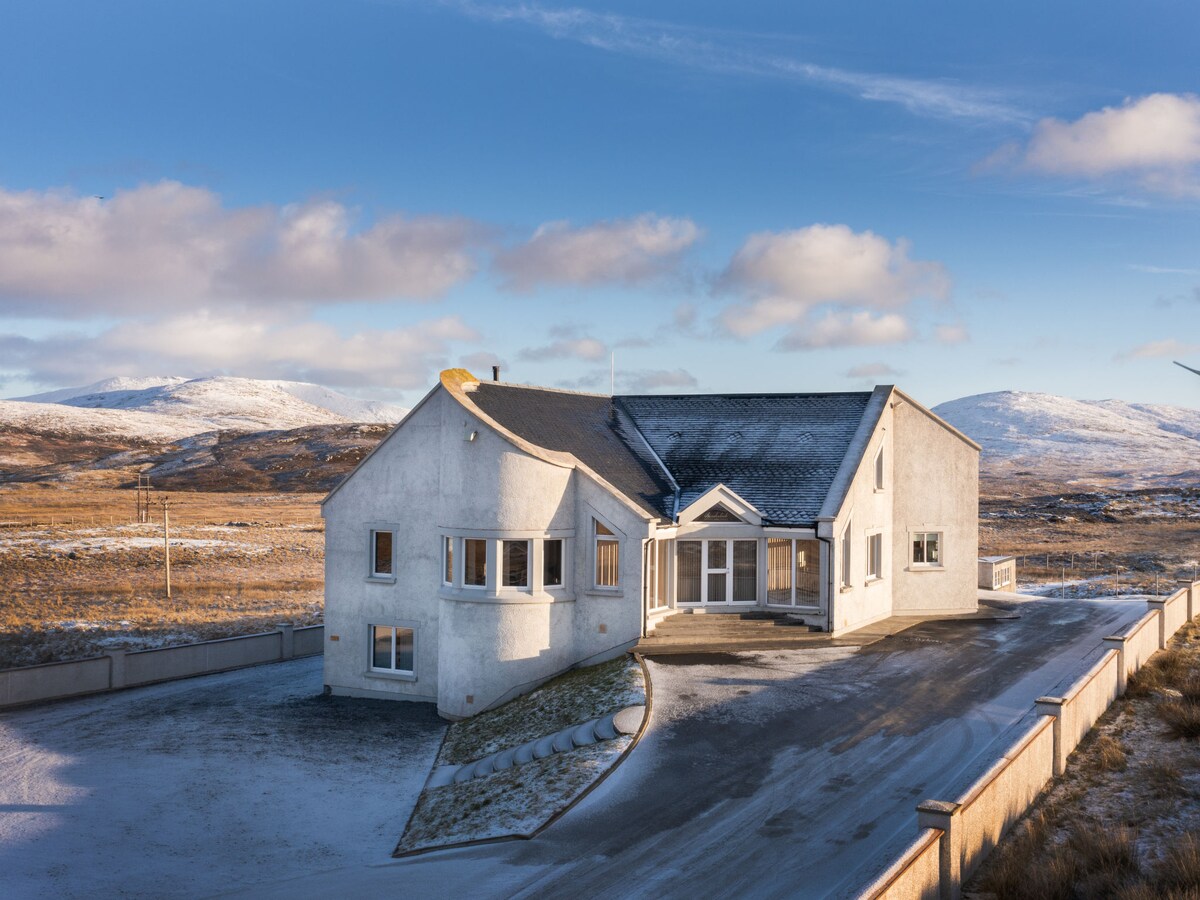 A modern, detached house is set against a backdrop of rolling hills and blue skies. The exterior features a clean design with large windows, providing ample natural light. A spacious driveway is visible, leading to the main entrance of the property.