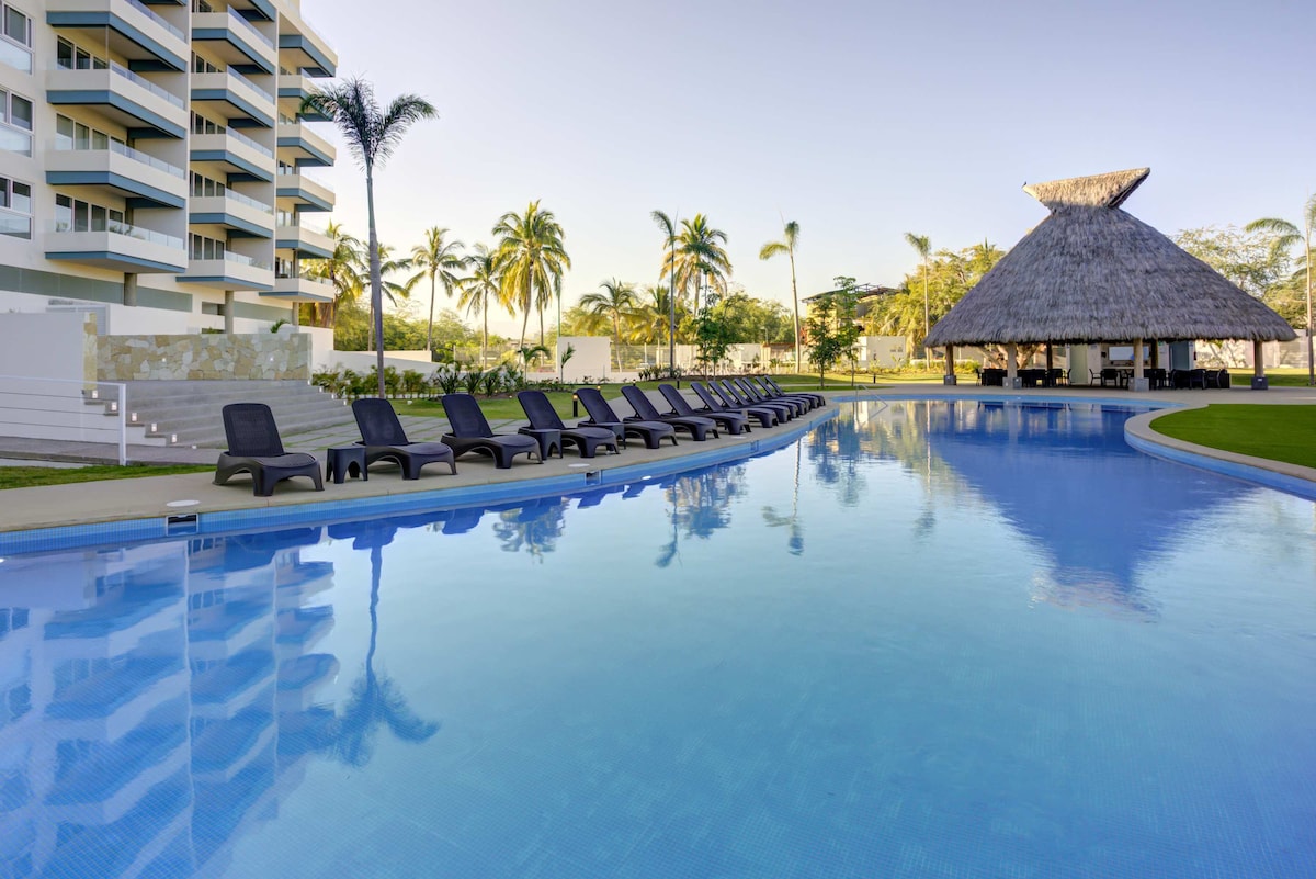 A large outdoor swimming pool features a tranquil expanse of blue water, lined with numerous lounge chairs. A thatched-roof palapa is visible in the background, surrounded by lush greenery and palm trees, creating a serene atmosphere.