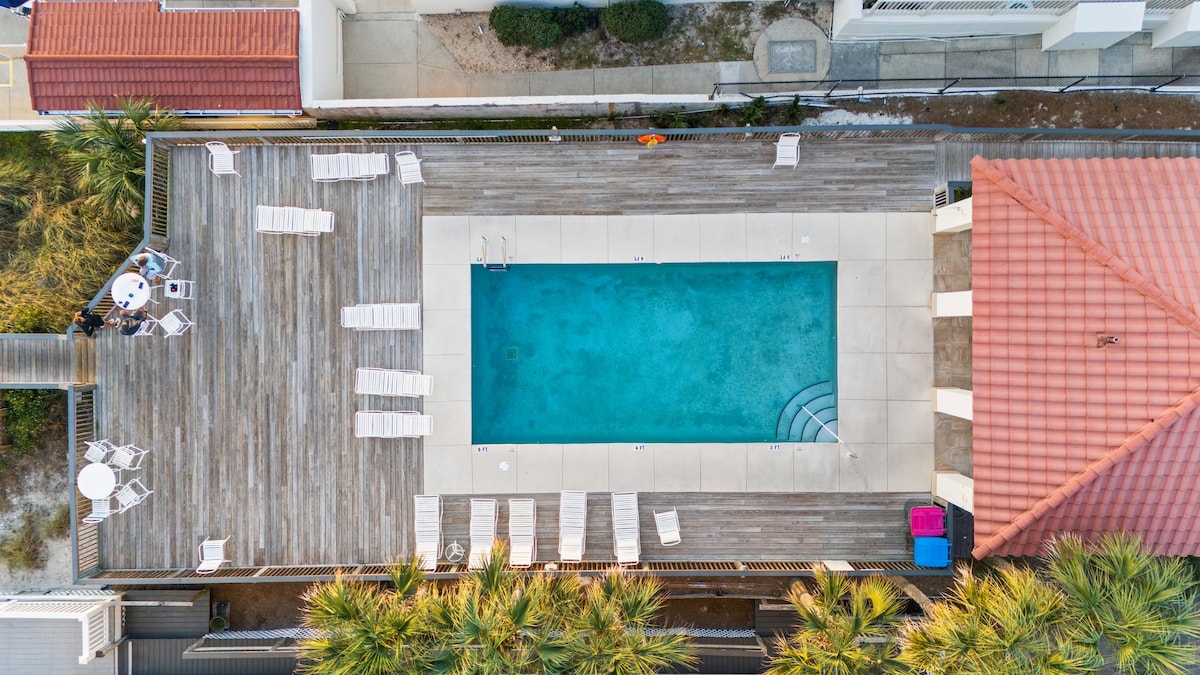 An aerial view of a beachside pool reveals a spacious rectangular area surrounded by wooden decking. Several lounge chairs are arranged around the pool, complemented by palm trees that provide a tropical atmosphere. The nearby building features a terracotta tiled roof.