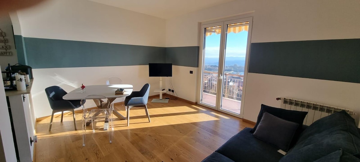A bright living area is presented with large glass doors that open to a panoramic view. A white dining table is surrounded by four chairs, while a comfortable sofa is positioned nearby. The room features light wooden flooring and striped wall accents in calming colors.