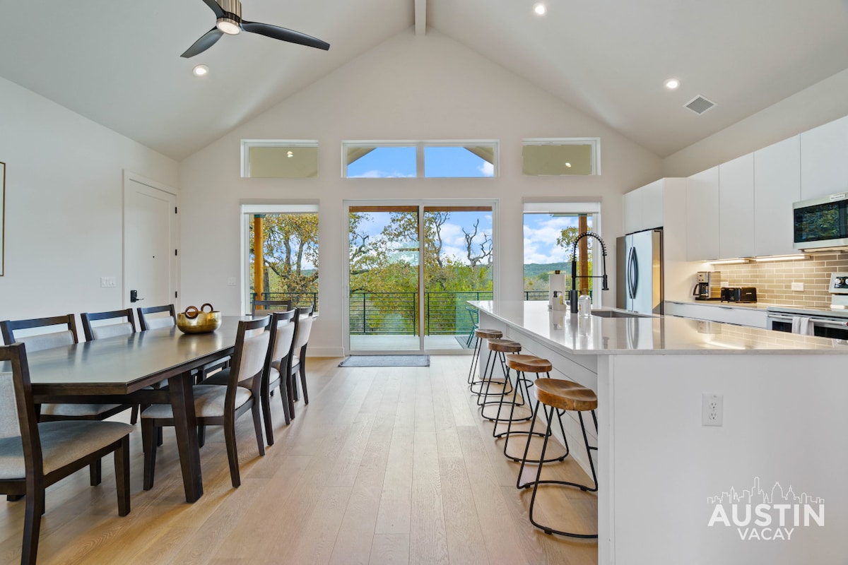 A spacious dining area is complemented by a long wooden table surrounded by chairs, with a view through large glass doors. Natural light streams in, highlighting the modern kitchen that features an island with seating and sleek appliances.