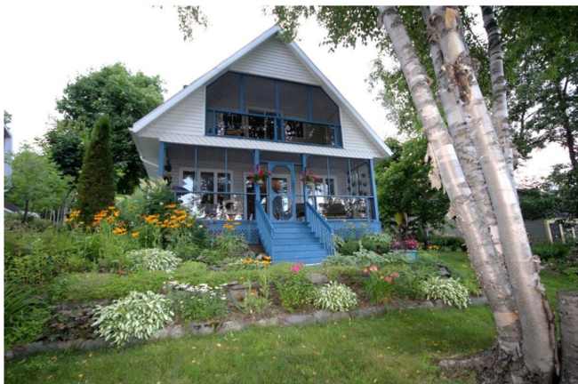A charming chalet exterior is shown, featuring a blue entrance and an extensive porch framed by vibrant flower beds. Surrounding trees add to the lush greenery, creating a serene outdoor environment. The building's architecture showcases a blend of traditional and inviting design elements.
