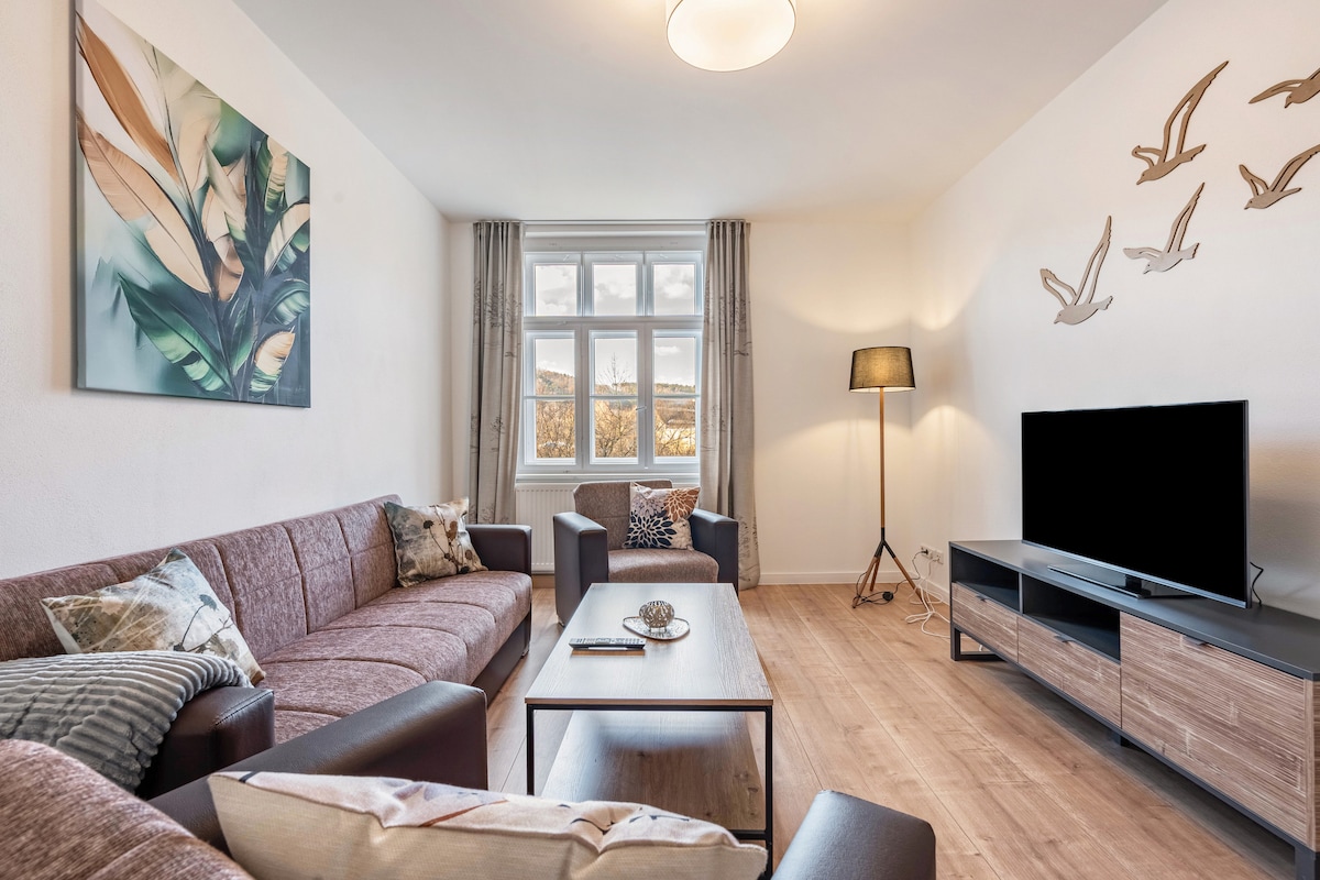 The living room features a cozy arrangement with a large sofa set and armchairs. A wooden table sits in the center, complemented by a decorative bowl. A flat-screen television is positioned on a media unit. Natural light filters through large windows, enhancing the serene atmosphere.