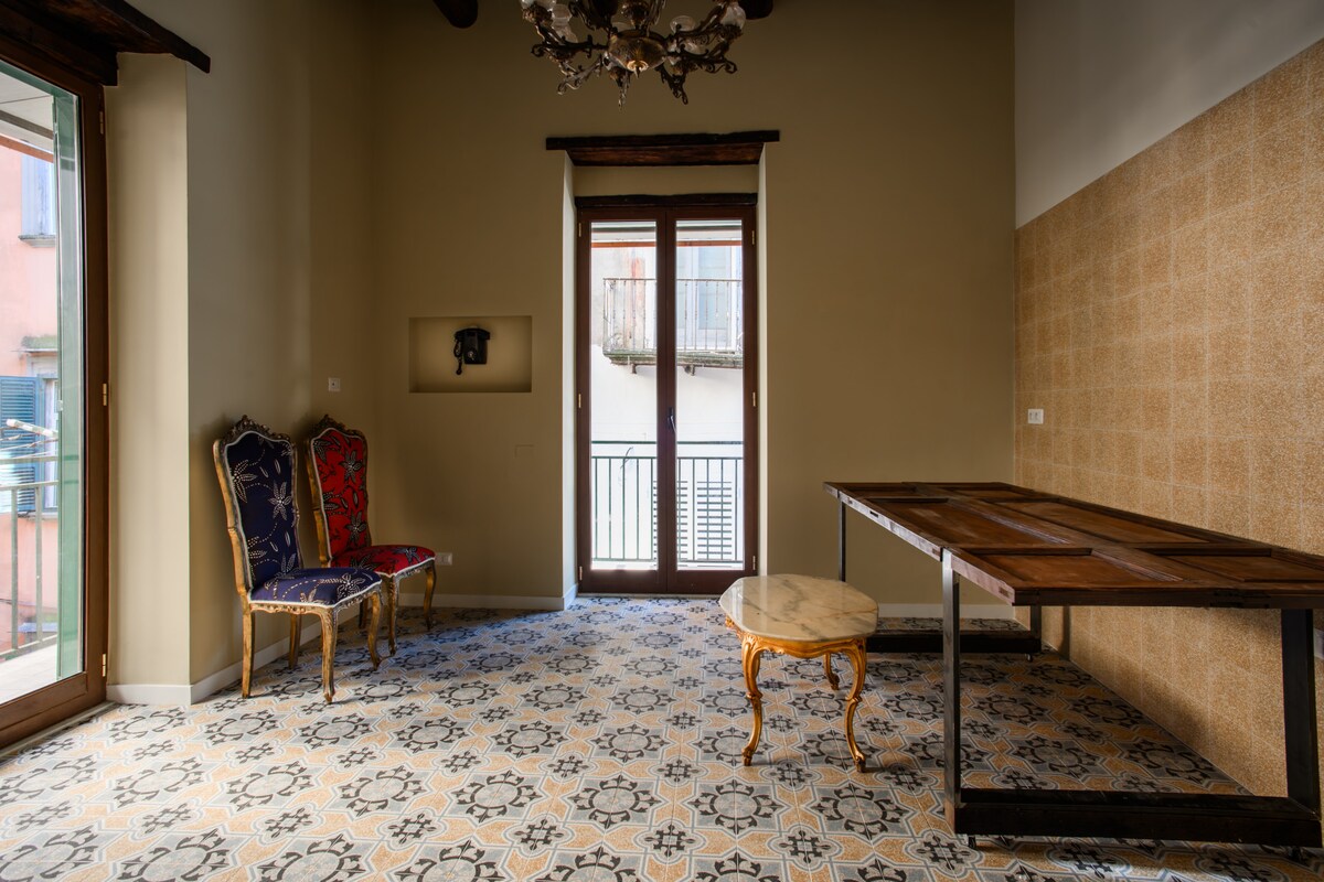 The spacious room features two ornate chairs, one in red and gold, the other in blue and gold, positioned near a balcony door. A small round table stands next to a long wooden table, with patterned tiles covering the floor.