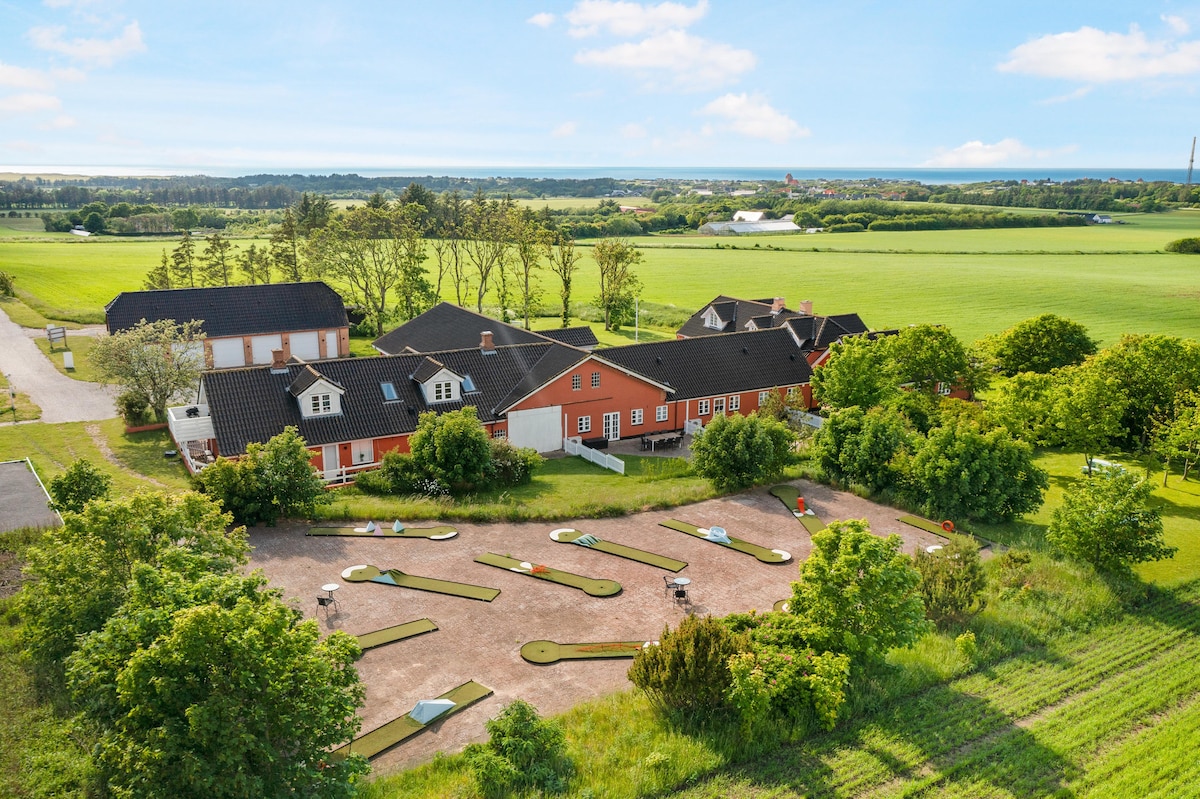 An overview of the property at Hawbakken showcases multiple holiday rentals surrounded by lush greenery. Mini-golf courses occupy the area in the foreground, while expansive fields extend towards the horizon, with the North Sea visible in the background.
