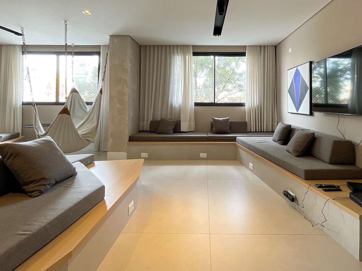 A modern living area features a large, minimalistic seating arrangement with gray cushions on inviting platforms. Natural light pours in through expansive windows framed by soft curtains, highlighting the spaciousness of the room. A contemporary artwork hangs on the wall opposite the seating, enhancing the overall aesthetic.