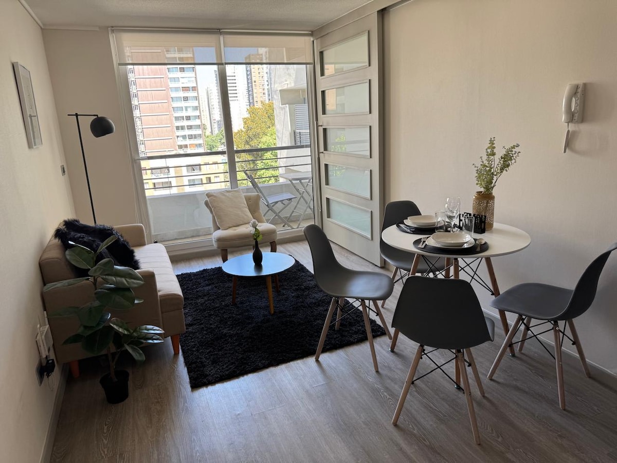 A bright living area is depicted, featuring a comfortable sofa, a small coffee table, and a dining setup for four. Natural light floods the space through large glass doors leading to a balcony. A soft rug adds warmth to the modern decor.