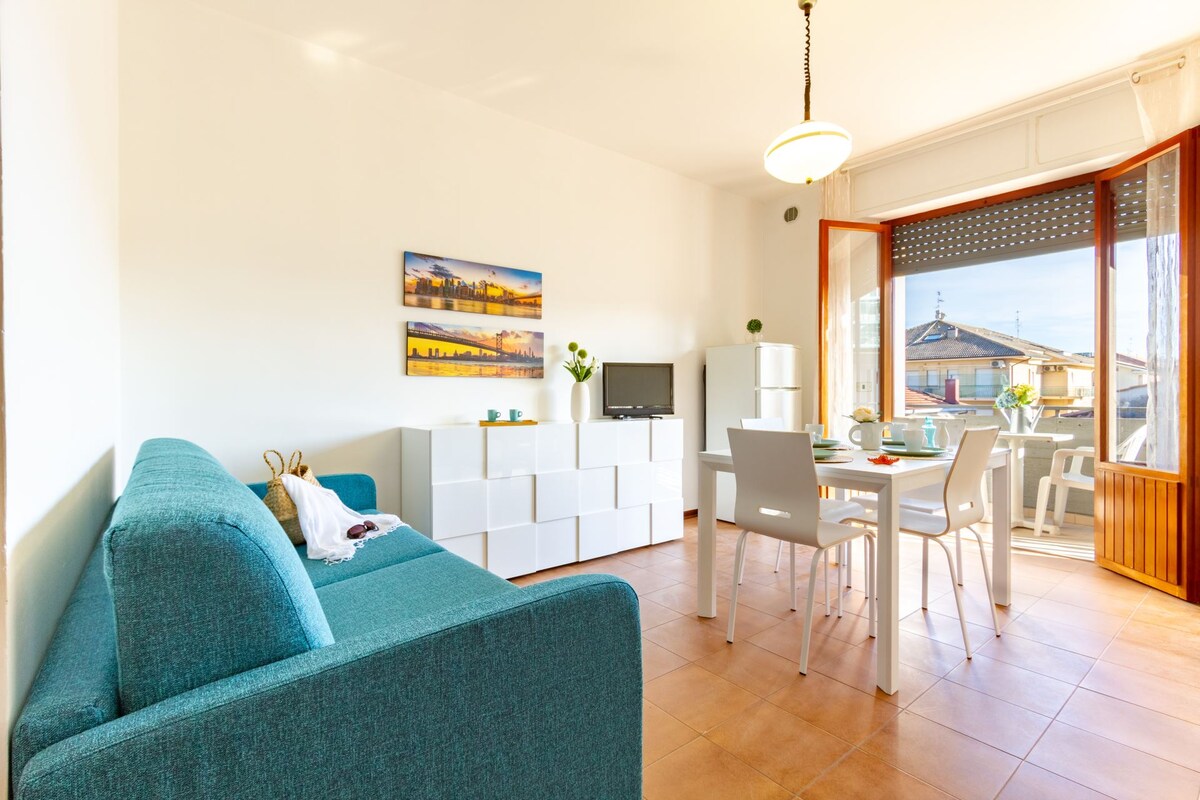 An open space living area features a teal sofa accompanied by white shelves and a television. A dining table with four chairs is positioned near large windows, offering access to the furnished balcony. Natural light fills the room, highlighting the tiled floor.
