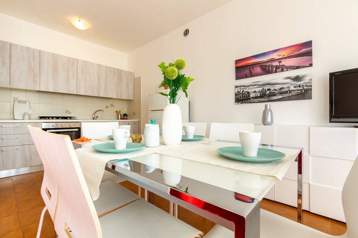 A modern open space kitchen and dining area is depicted, featuring a glass dining table set with plates and cups. White chairs surround the table, and a kitchen area with light wood cabinetry is visible in the background. A colorful artwork is displayed on the wall.