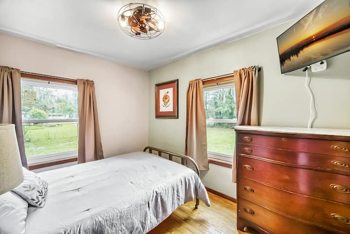 Encantador dormitorio con ventanas grandes, una cómoda cama, cómoda de estilo antiguo y un televisor montado en la pared, perfecto para una estancia tranquila.