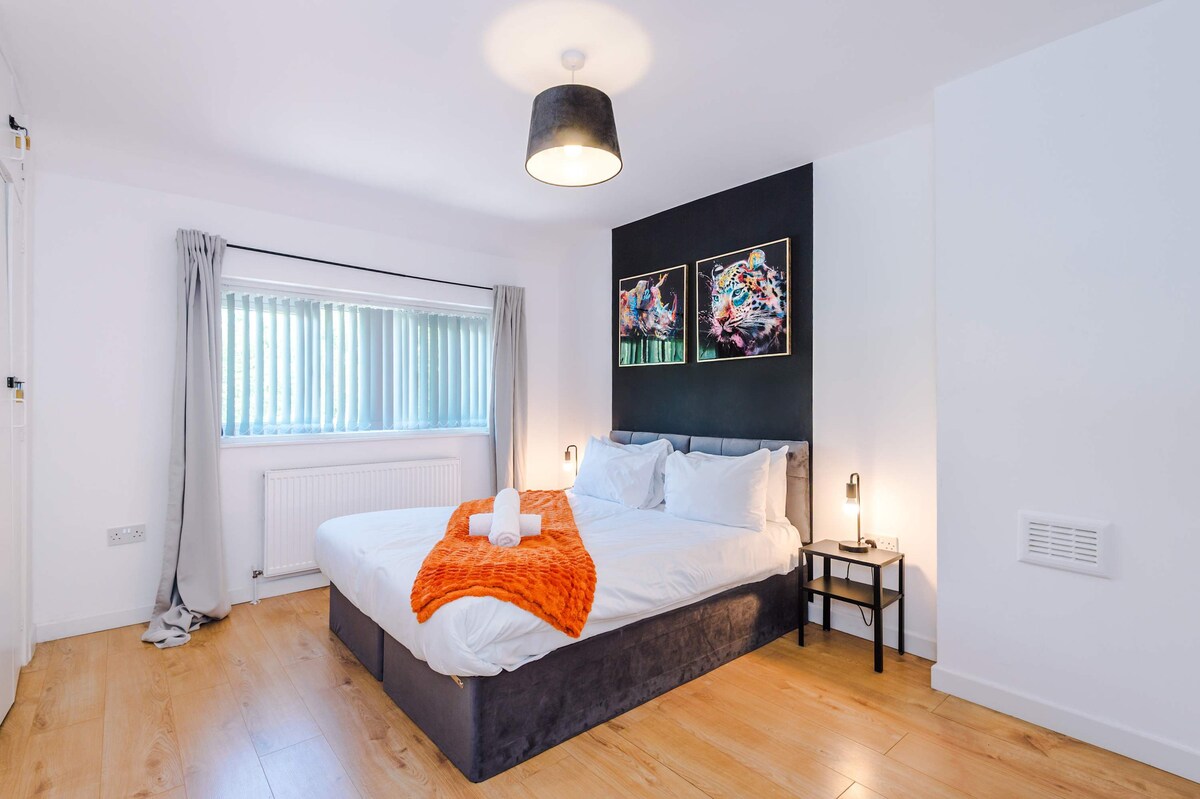 A spacious sleeping area features a large bed with white linens and a vibrant orange throw, complemented by two decorative pillows. Framed artwork adorns the wall above the bed, and natural light filters through vertical blinds on the window.