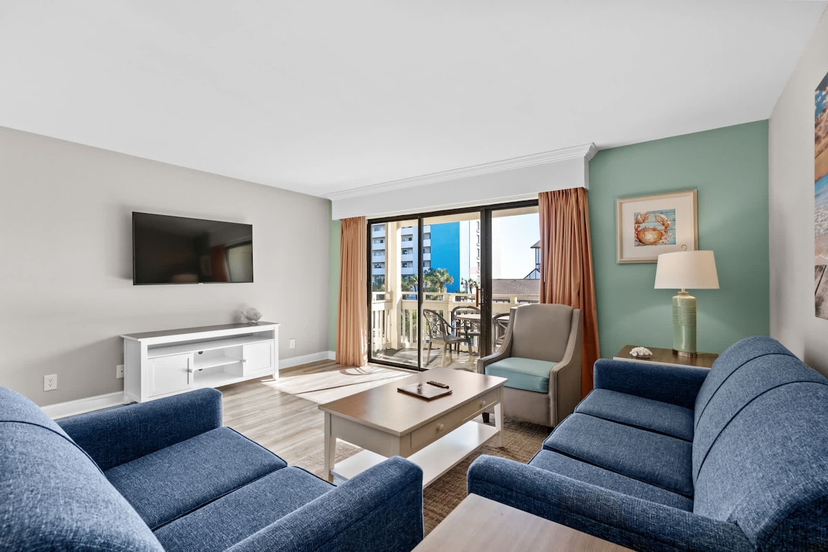 An open living area features a large flat-screen TV mounted on a light-colored wall. Two blue sofas face a coffee table, while natural light floods in through sliding glass doors leading to a balcony. Light-toned wood flooring complements the soft color palette.