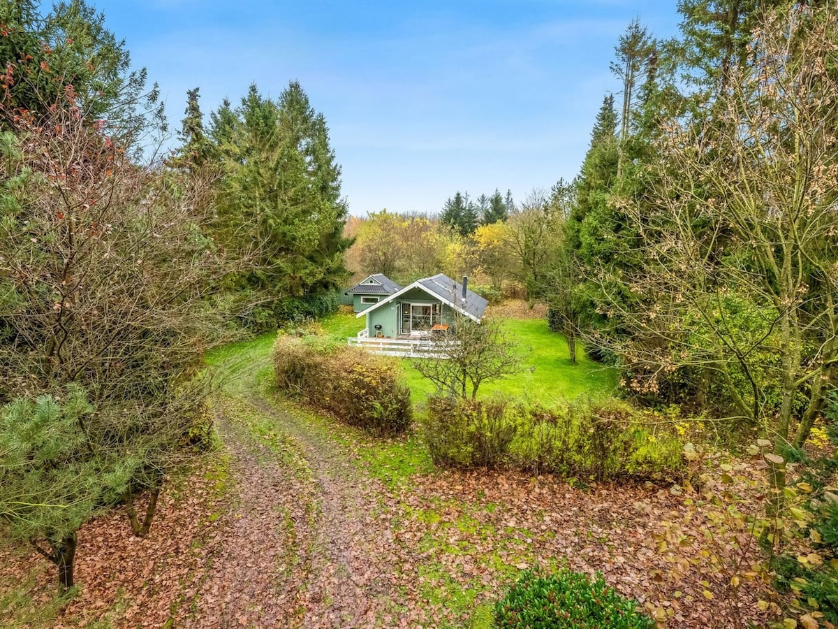 A single-family house is set amidst greenery, surrounded by trees and bushes. A winding path leads to the entrance, which features a porch. The natural landscape is adorned with fallen leaves, suggesting a tranquil and serene outdoor environment.