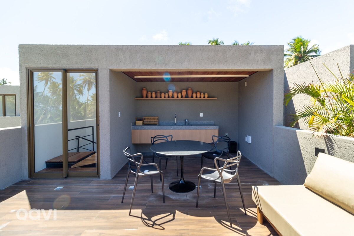 An inviting outdoor terrace features a sleek wooden deck, complemented by a dining table with chairs. A minimalist counter with decorative pottery adds a touch of style, while lush greenery peeks from the edges, creating a serene space for relaxation.