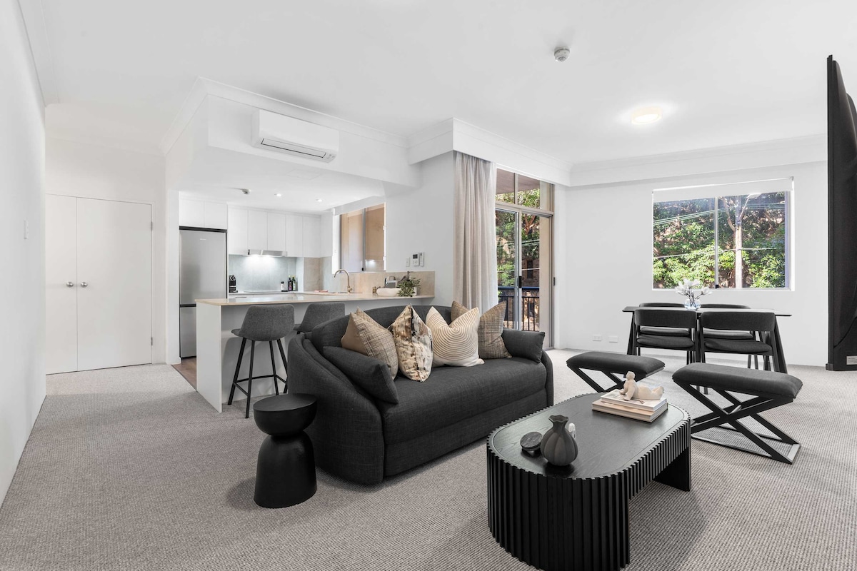 An open-plan living area is depicted, showcasing a comfortable black sofa adorned with decorative pillows. A coffee table with a textured design complements the seating. To the left, a dining area with a dark table and chairs is visible, while a modern kitchen can be seen in the background.