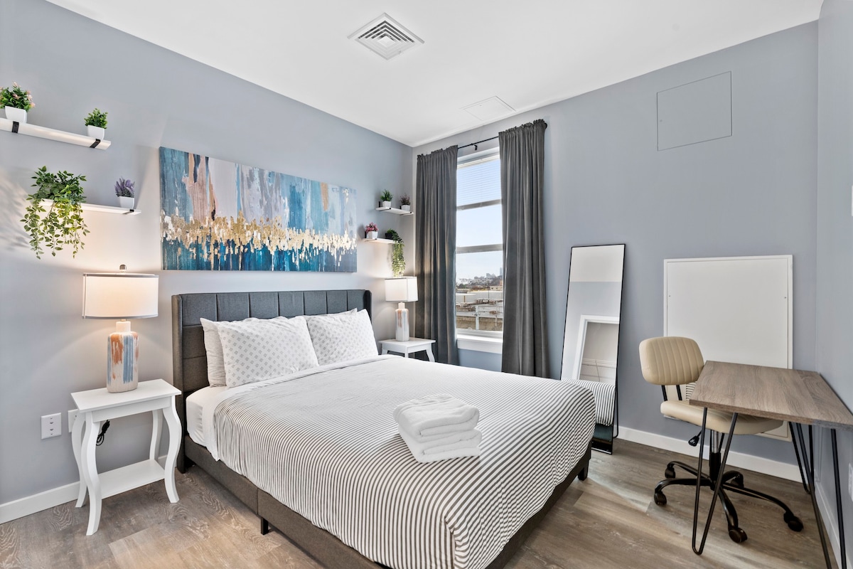 A well-lit bedroom features a spacious bed with striped bedding and two bedside tables, each with a lamp. A desk and chair are positioned near the window, complemented by wall shelves with plants and artwork above the bed. A mirror enhances the room's dimensions.