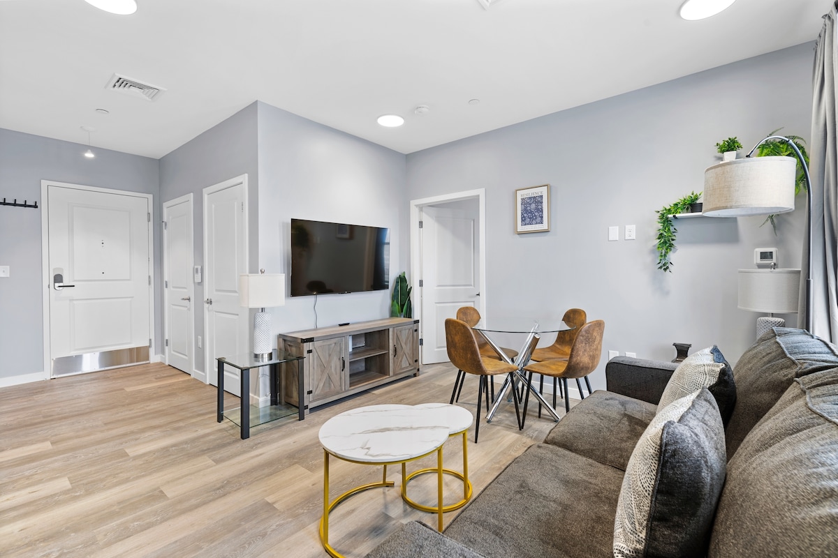 A modern living area features a comfortable sectional sofa alongside a coffee table. A dining table with four chairs is positioned near a window. A wall-mounted television and decorative shelving add to the functional aesthetic, while natural light filters through the curtains.