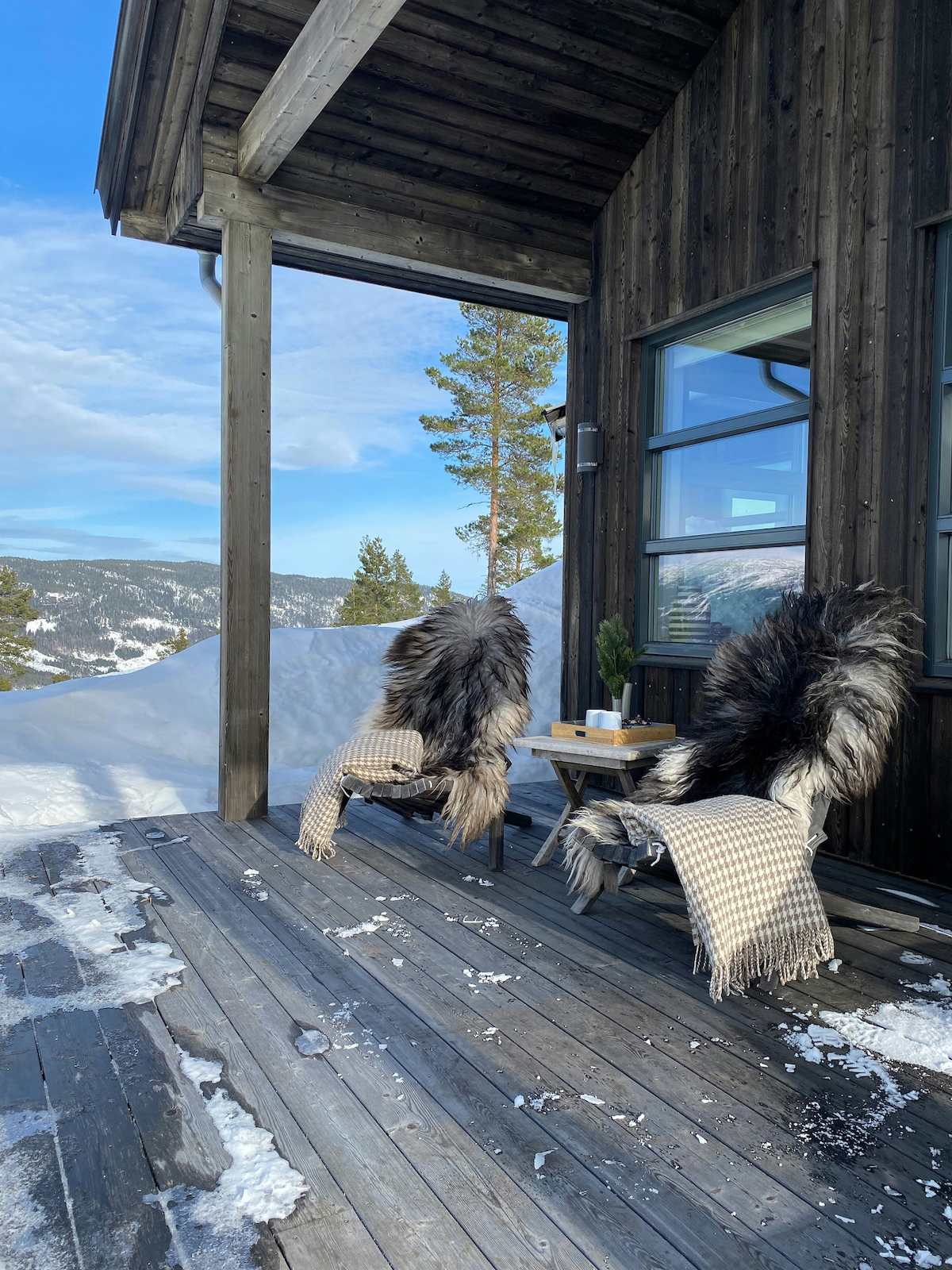 A terrace features two comfortable chairs adorned with soft, fur throws, positioned beside a small wooden table. Snow blankets the ground, contrasting with the dark wood of the cabin. A serene view of mountains and trees is visible in the background under a clear blue sky.