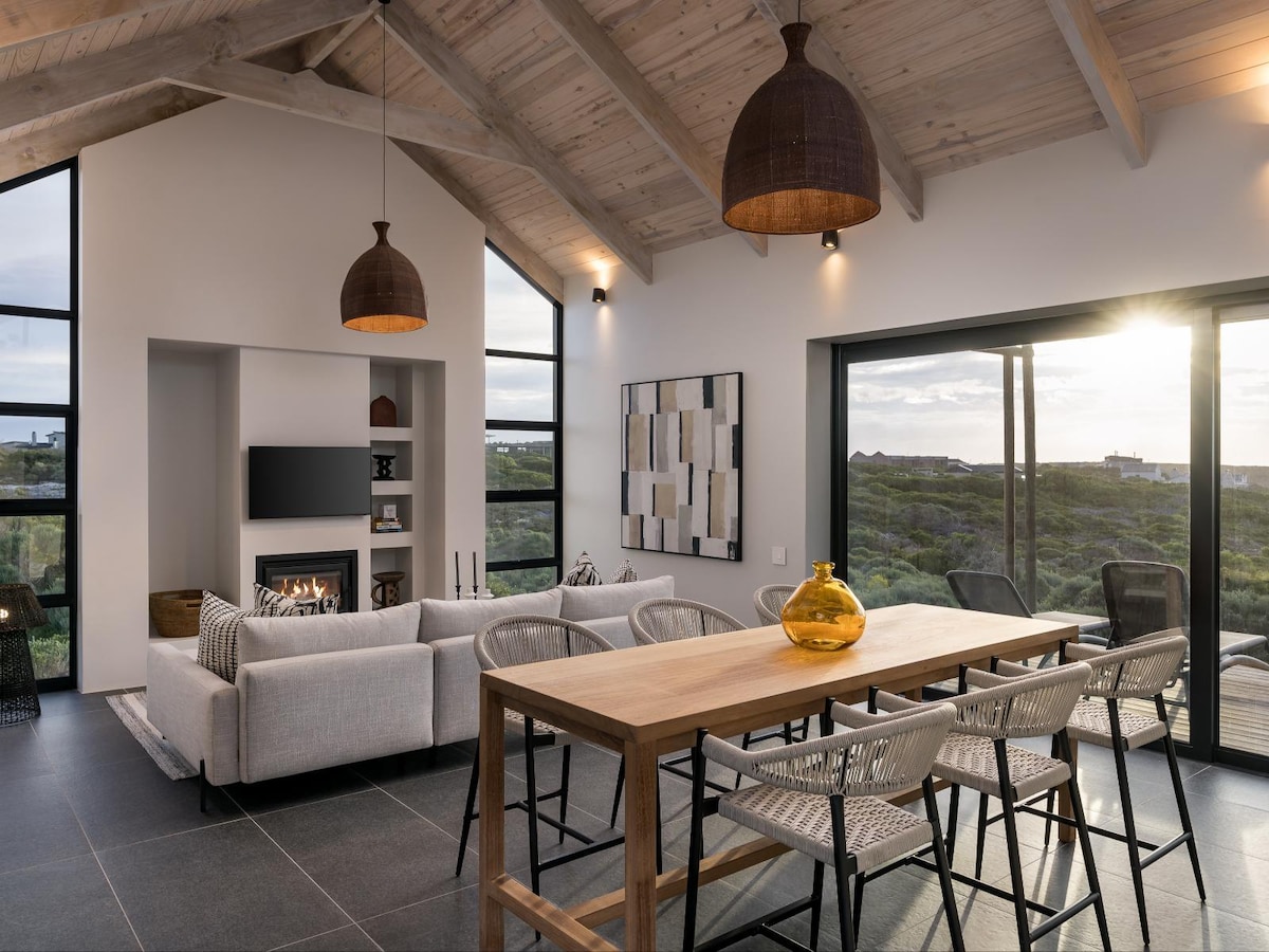 A spacious living area features a high, wooden-beamed ceiling and large windows showcasing outdoor views. A modern gray sofa is arranged with decorative cushions, while a stylish dining table is accompanied by several woven chairs. An inviting fire is visually framed in the contemporary fireplace.