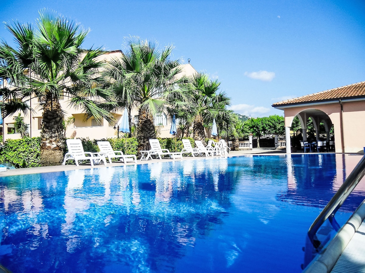 A clear blue swimming pool is surrounded by palm trees, with white lounge chairs arranged neatly along its edge. The vibrant greenery and the building in the background create a serene atmosphere, inviting relaxation under the sun.