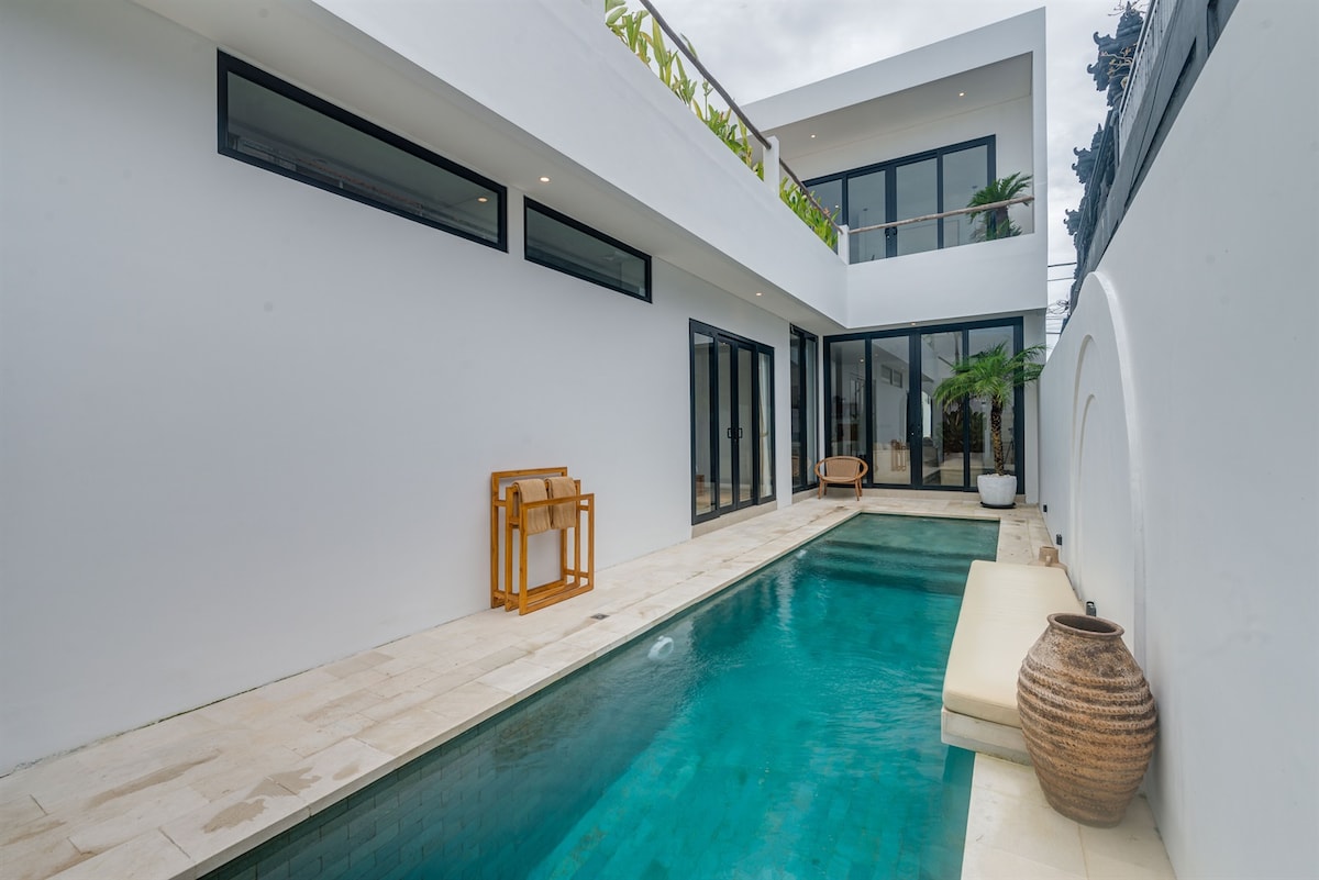 A modern villa features a serene private pool, bordered by minimalist stone tiles. Glass doors offer seamless access to the indoor area, which is complemented by lush greenery. A wooden bench and a sun lounger provide inviting spaces to relax by the water.